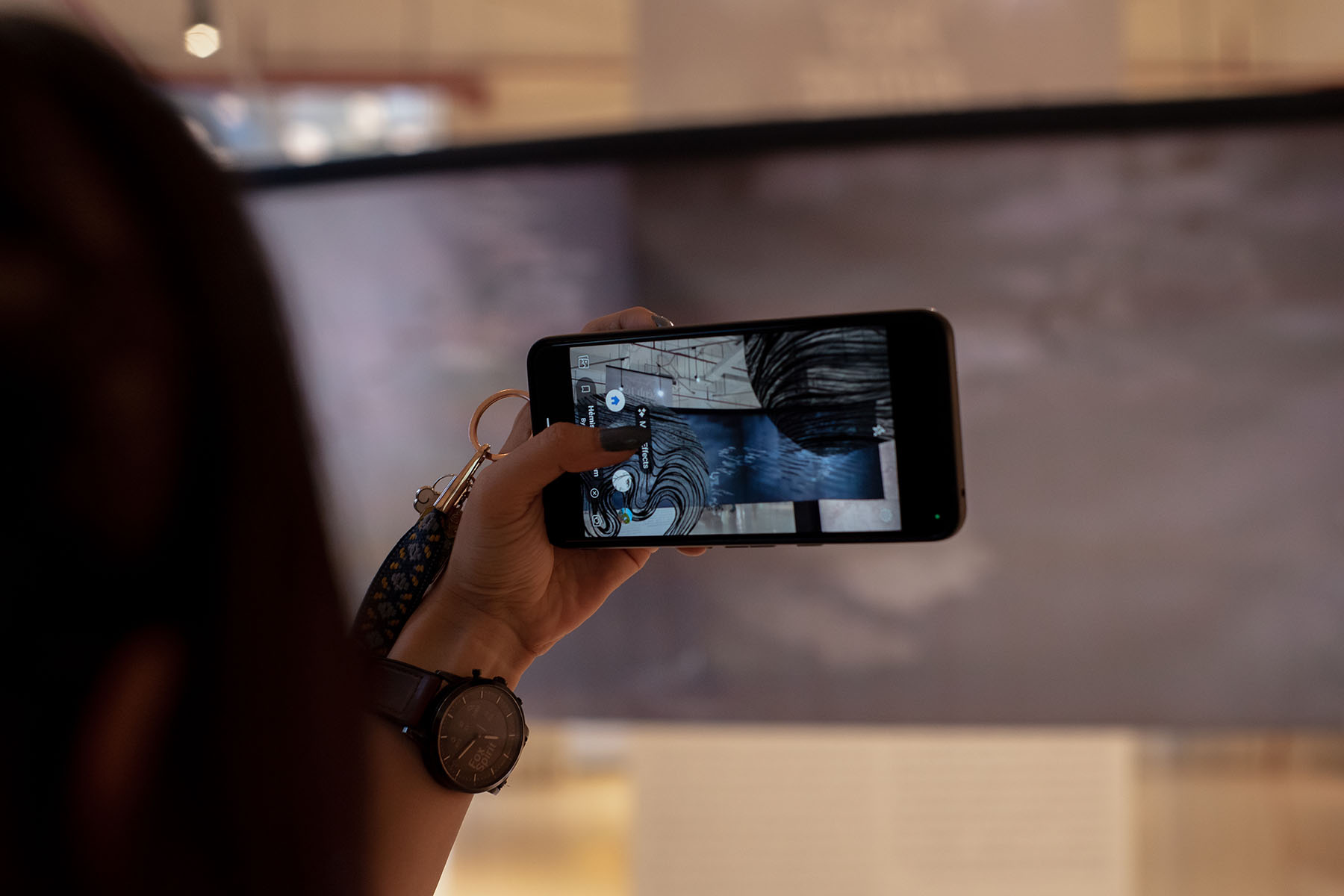 A woman scanning augmented reality installation with a smartphone. Visible smartphone augmented abstract shapes on the screen. Printed dark canvas in the background.