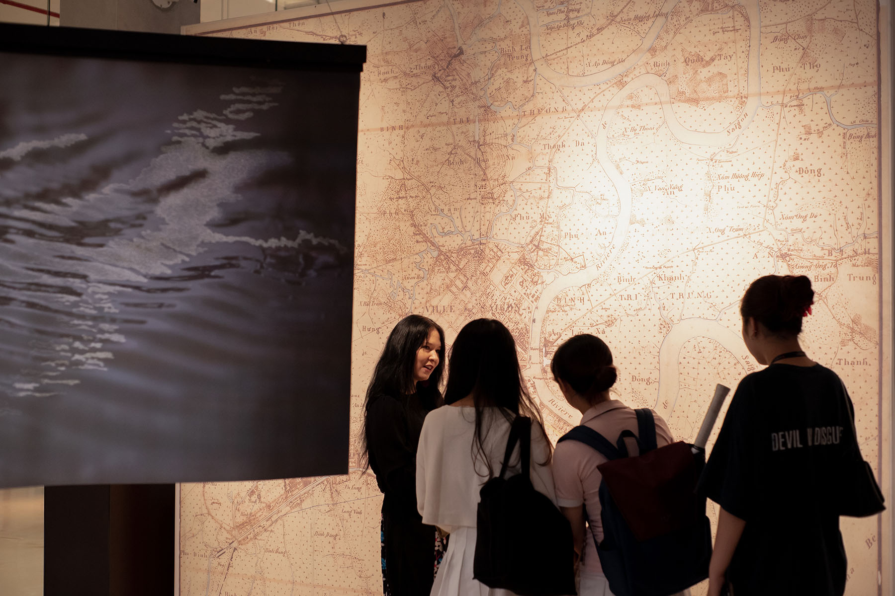 A group of four people standing in front of the big historical map of Saigon. A woman is explaining the map; three girls are listening. Another printed work with water shapes visible on the left.