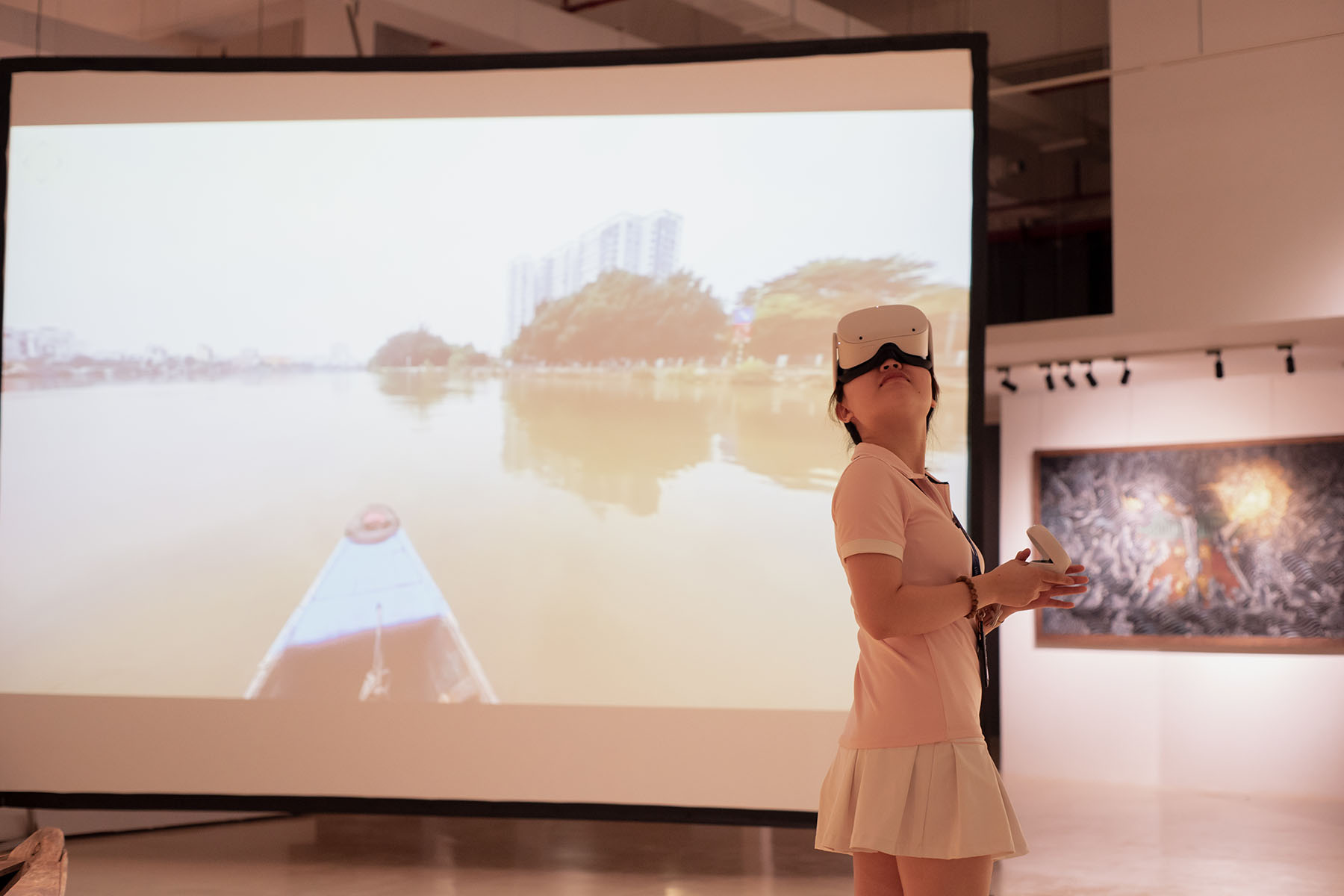 A girl in the VR Oculus Go headset is standing in front of the screen. There is a wooden boat on the water on the screen. The art gallery's white surroundings in the background 