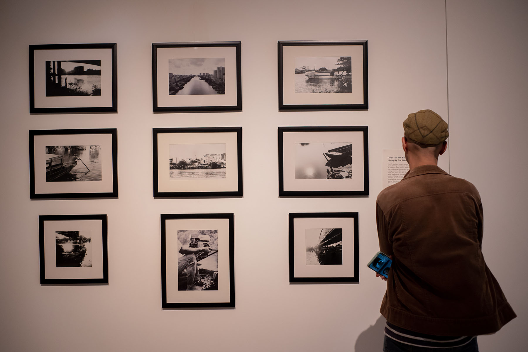 A man watching photographs and reading a description. Nine photos organized in three rows. A printed description on the right, in front of the man. Photos depicting Kenh Te and scenes from living close to the water.